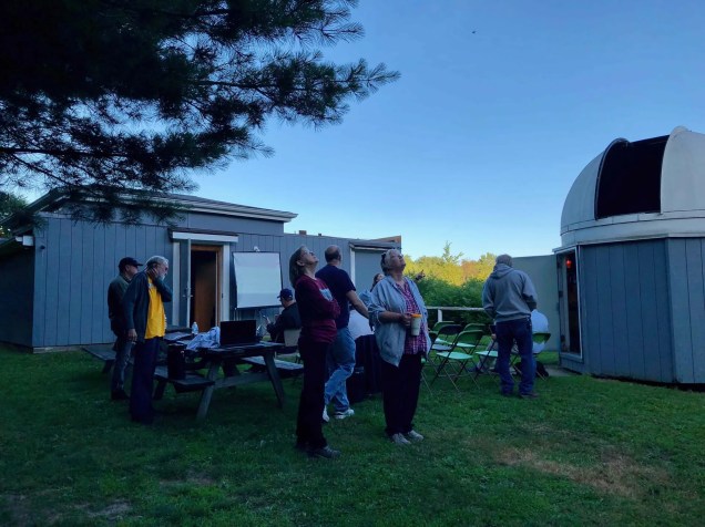 Several club members staning between two observatory buildings at their Indian Hill Observatory site. Rear, left, is a roll-off roof type building, and on the right is a traditional dome-topped enclosure.